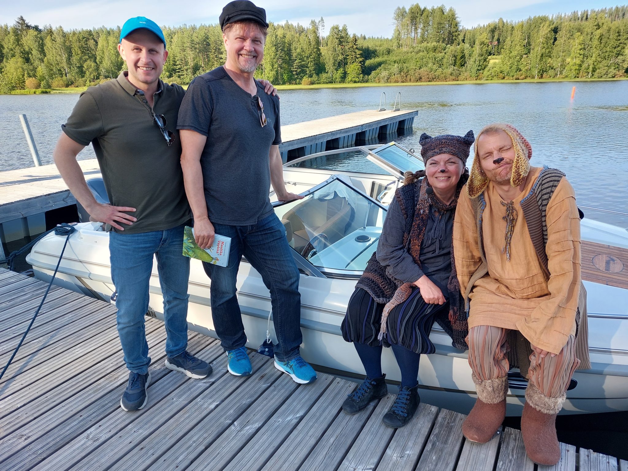 Neljä ihmistä poseeraa telakoidun veneen edessä, taustalla rauhallinen järvi ja metsä. Kaksi heistä on pukeutunut rennosti, toinen siniseen ja toinen mustaan lippikseen, kun taas kaksi on pukeutunut värikkäisiin asuihin, mukaan lukien neulottu hattu ja tunika.