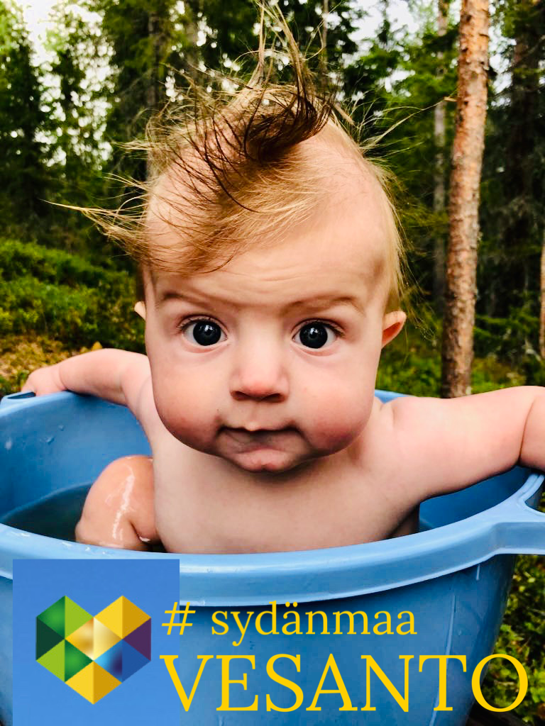 Piikkihiuksinen vauva kylpee sinisessä ammeessa ulkona puiden ympäröimänä. Kuvassa teksti "# sydänmaa VESANTO" ja sen kulmassa monivärinen geometrinen logo.