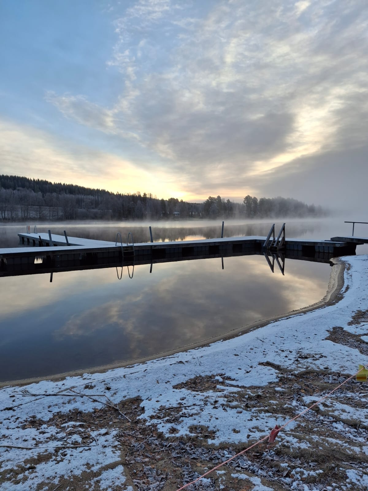 Rauhallinen järvenrantamaisema, jossa on lumipölyinen ranta ja pitkä laituri, joka ulottuu tyynelle vedelle. Järvestä nousee sumua, ja taustaa reunustavat tiheät puut, kun taivas vaihtuu aamunkoitosta päivään.