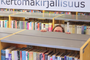 Kuvassa opastekyltti kertomakirjallisuus. nainen kurkistaa kirjahyllyn yli.